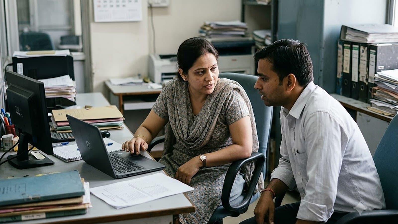 A manager marking a printed rating sheet at a desk in a branch office, a colleague reviewing the same sheet beside her